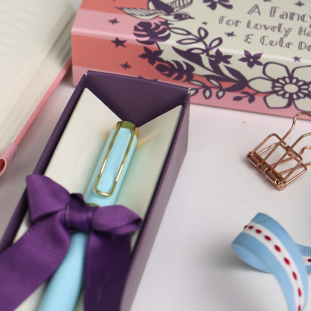 Blue pen with purple ribbon in a box, surrounded by stationery items on a white surface.