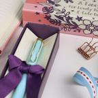 Blue pen with purple ribbon in a box, surrounded by stationery items on a white surface.
