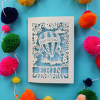 A paper cut bon voyage card, lased cut from cream card with a light blue paper backing. The card has a hot air balloon with "bon voyage" and a name underneath