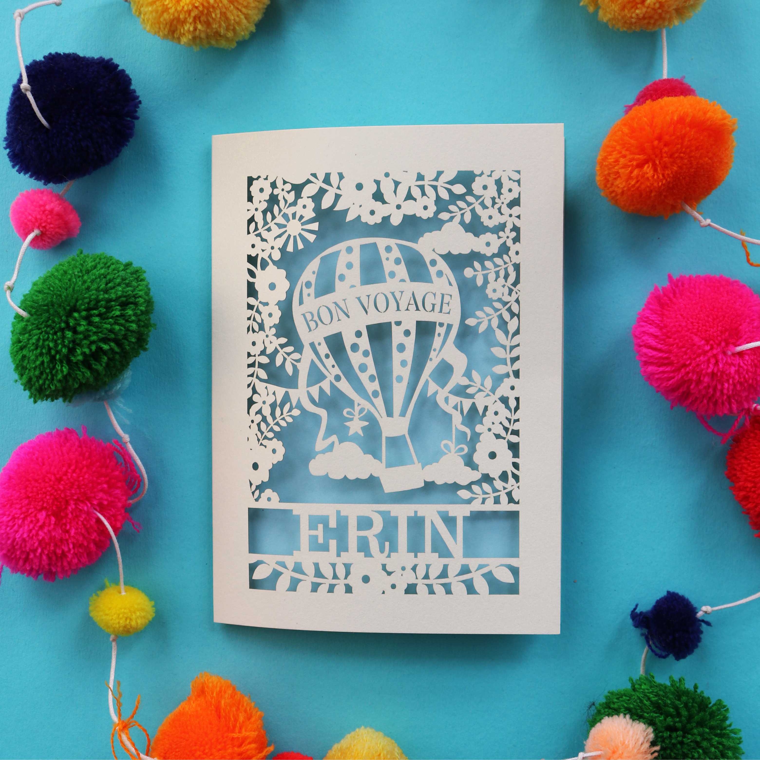 A paper cut bon voyage card, lased cut from cream card with a light blue paper backing. The card has a hot air balloon with "bon voyage" and a name underneath