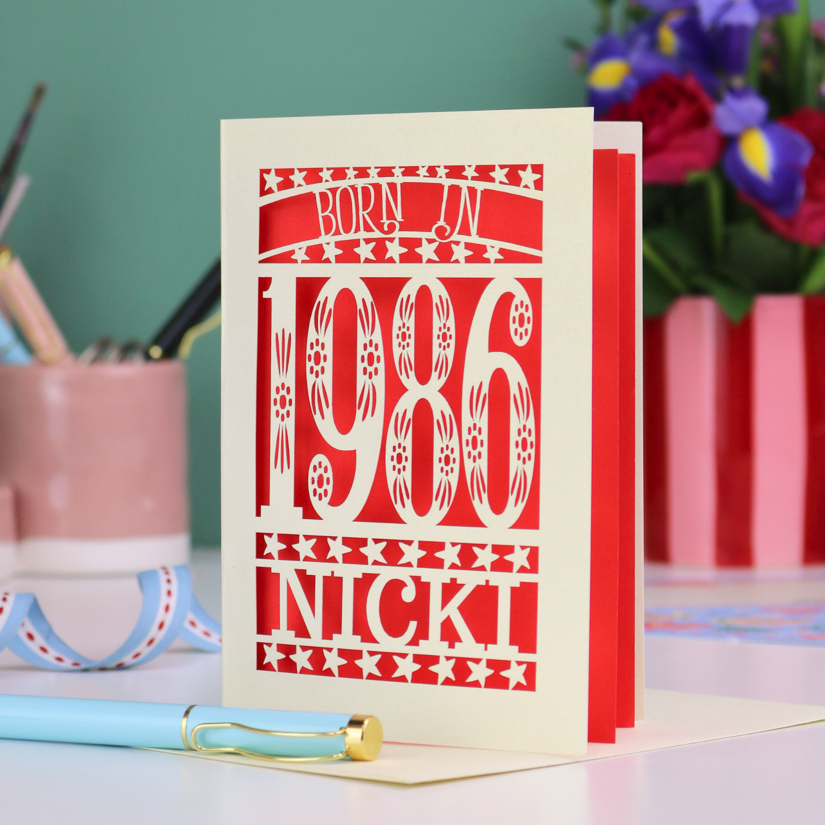 Greeting card with 'Born in 1986 Nicki' design on a desk with stationery items.