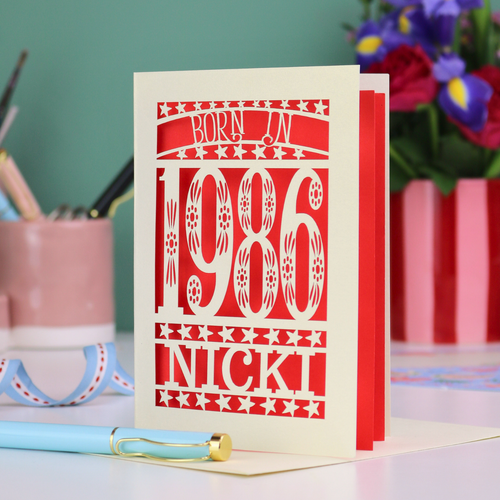 Greeting card with 'Born in 1986 Nicki' design on a desk with stationery items.