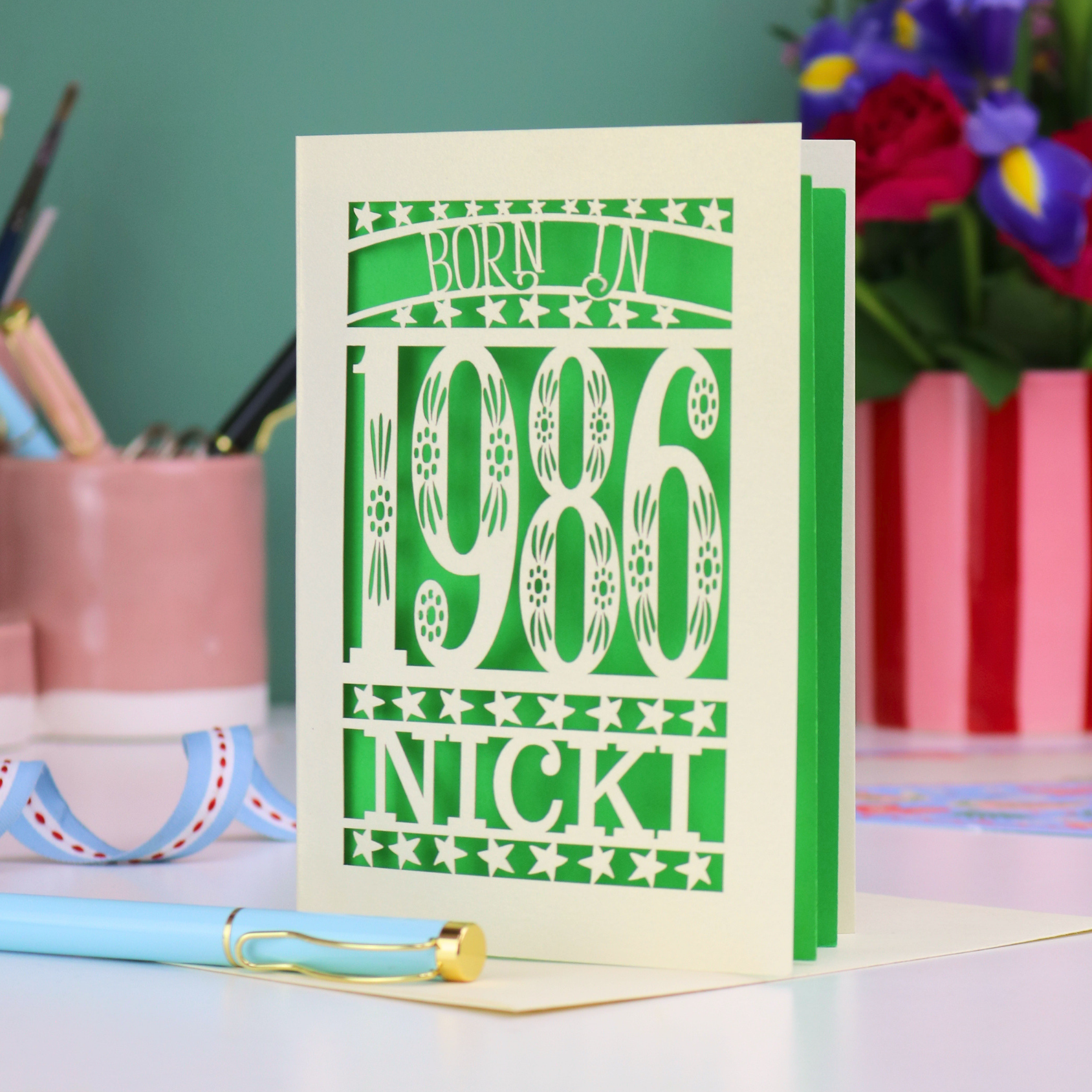 Card with 'Born in 1986 Nicki' design on a desk with stationery items.