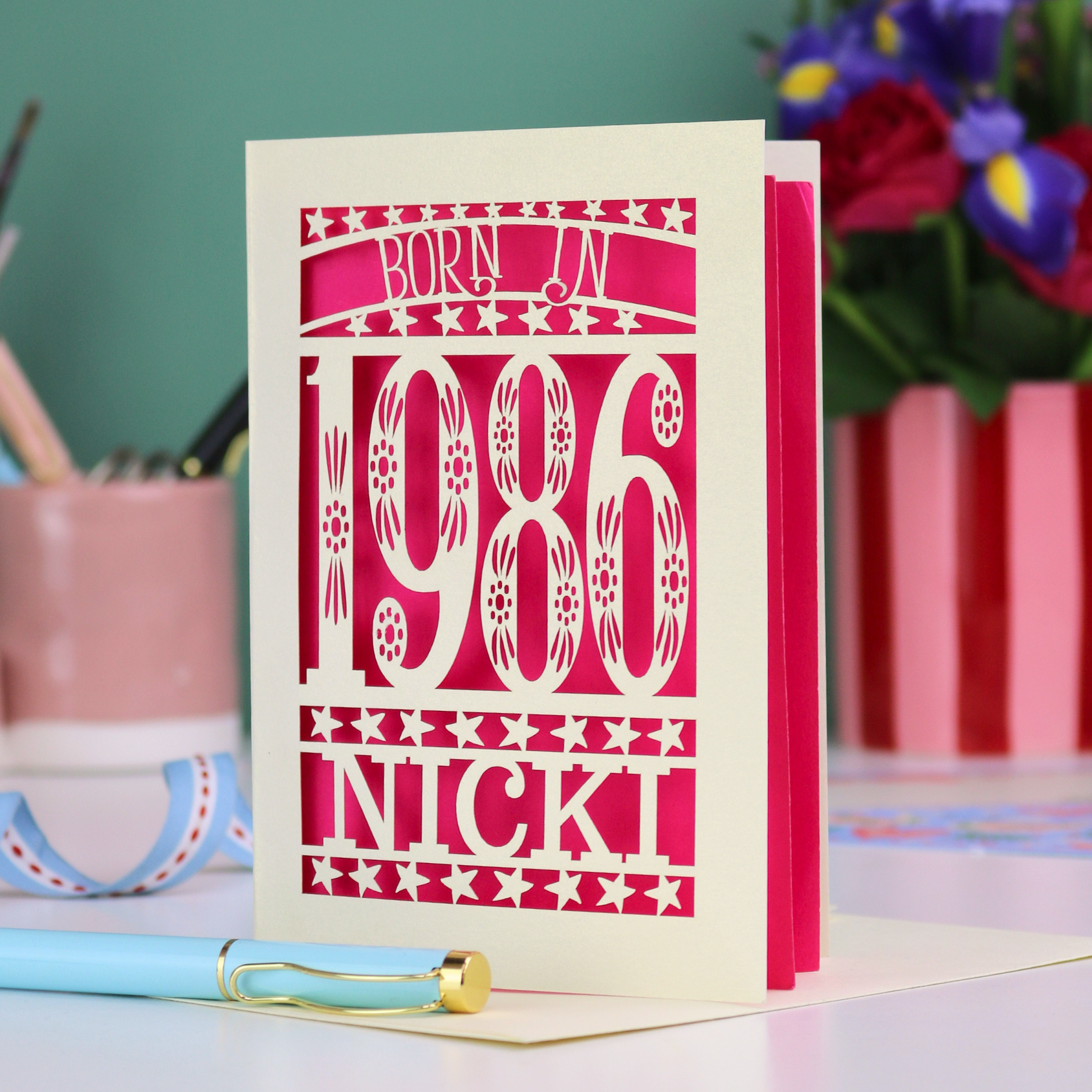 Personalized birthday card with 'Born in 1986 Nicki' design on a desk with flowers and stationery.