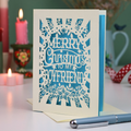 A laser-cut boyfriend Christmas card with the text 'Merry Christmas to my Boyfriend' in cream cut out lettering over a blue background, displayed on a tabletop with candles and a pen in a softly blurred setting.