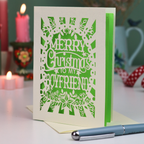 A laser-cut Christmas card for boyfriend, with the text 'Merry Christmas to my Boyfriend' in cream cut out lettering over a green background, displayed on a tabletop with candles and a pen in a softly blurred setting.