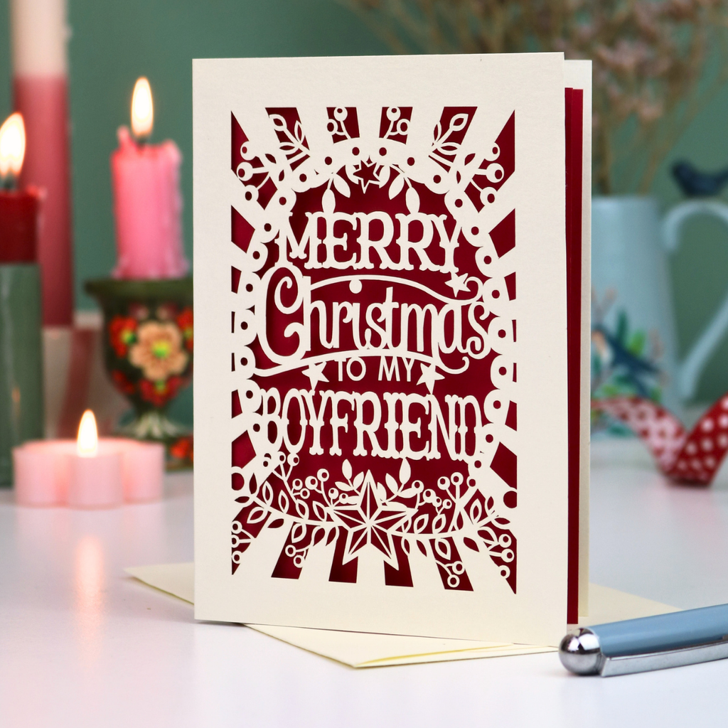 A laser-cut Christmas boyfriend card with the text 'Merry Christmas to my Boyfriend' in cream cut out lettering over a dark red background, displayed on a tabletop with candles and a pen in a softly blurred setting.