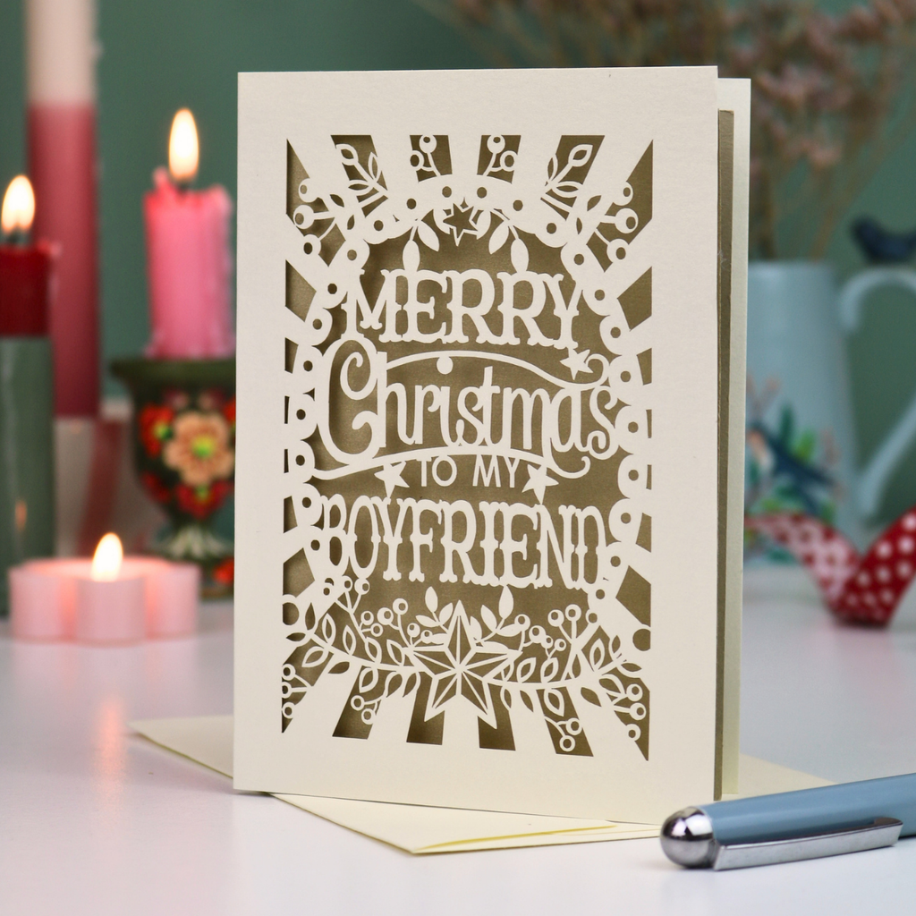 A laser-cut boyfriend Christmas card with the text 'Merry Christmas to my Boyfriend' in cream cut out lettering over a gold leaf background, displayed on a tabletop with candles and a pen in a softly blurred setting.
