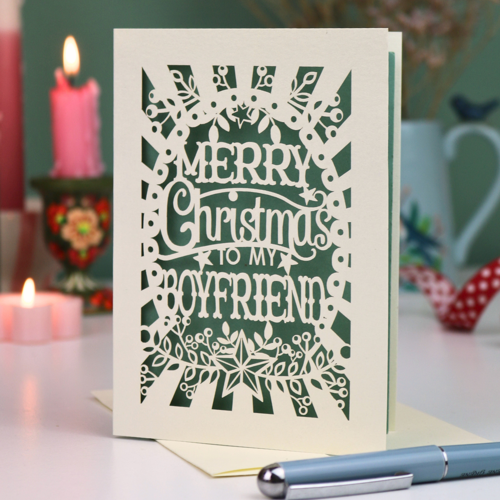 A laser-cut boyfriend Christmas card with the text 'Merry Christmas to my Boyfriend' in cream cut out lettering over a sage green background, displayed on a tabletop with candles and a pen in a softly blurred setting.