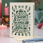 A laser-cut boyfriend Christmas card with the text 'Merry Christmas to my Boyfriend' in cream cut out lettering over a sage green background, displayed on a tabletop with candles and a pen in a softly blurred setting.