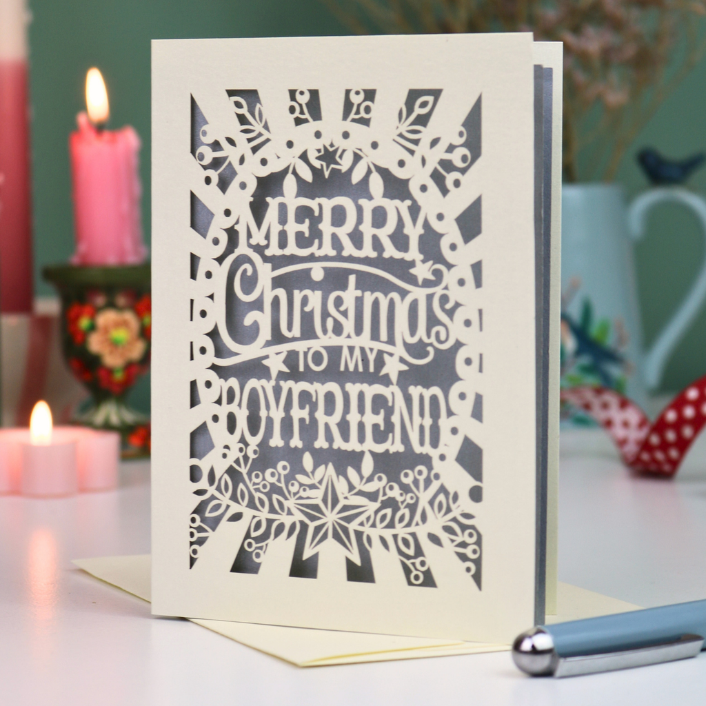 A laser-cut boyfriend Christmas card with the text 'Merry Christmas to my Boyfriend' in cream cut out lettering over a silver background, displayed on a tabletop with candles and a pen in a softly blurred setting.