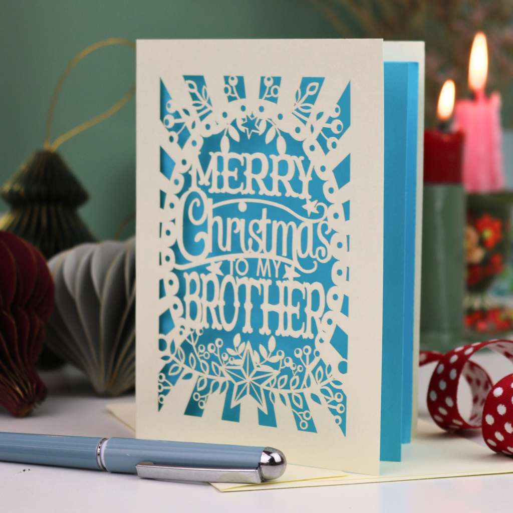 A handmade laser-cut cream and bright blue Christmas card reading 'MERRY Christmas TO MY BROTHER'. The card is displayed on a tabletop with a blue pen in the foreground and a lit candle in the background, creating a cosy holiday atmosphere.
