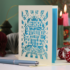 A handmade laser-cut cream and bright blue Christmas card reading 'MERRY Christmas TO MY BROTHER'. The card is displayed on a tabletop with a blue pen in the foreground and a lit candle in the background, creating a cosy holiday atmosphere.