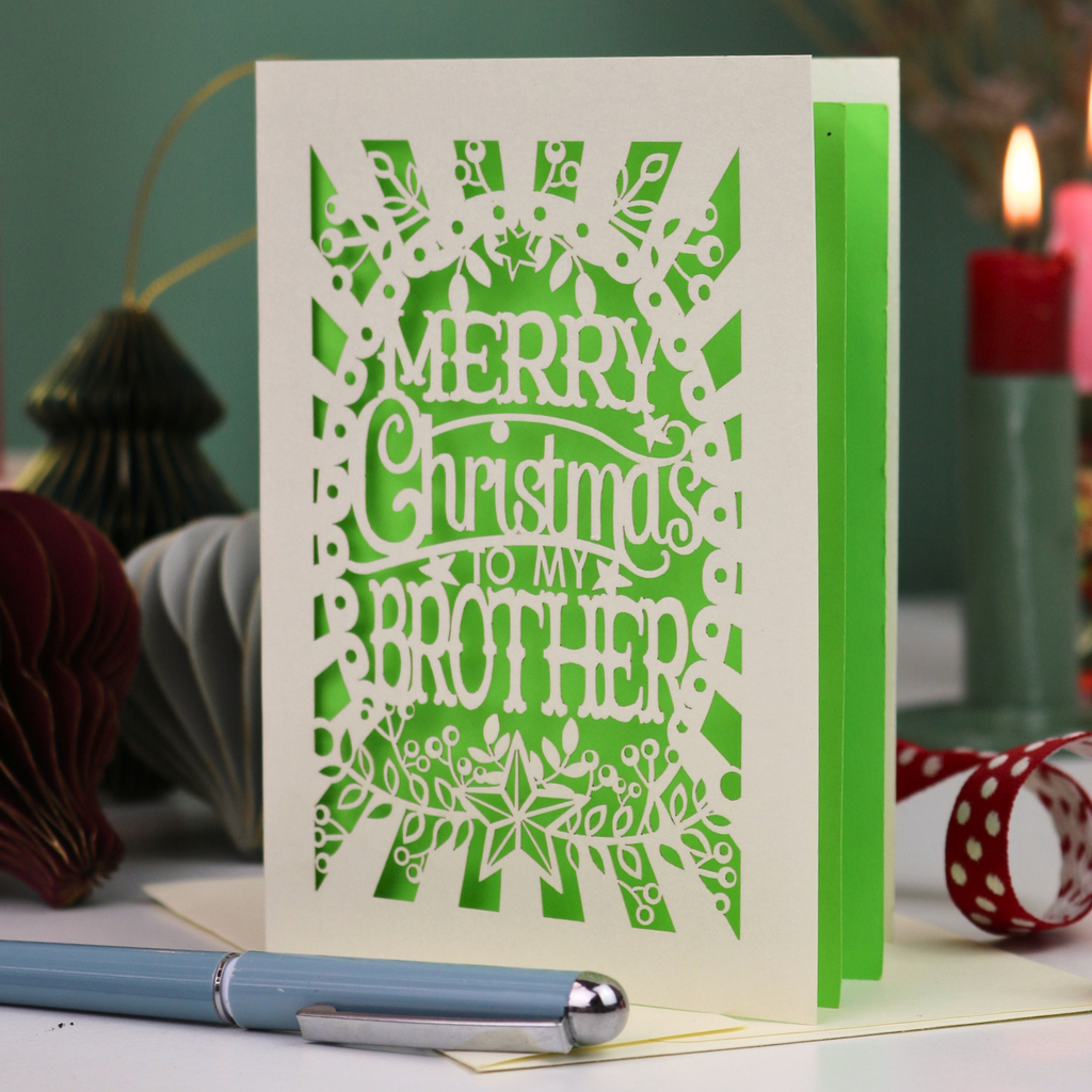 A handmade laser-cut Christmas card reading 'MERRY Christmas TO MY BROTHER'. The card is displayed on a tabletop with a blue pen in the foreground and a lit candle in the background, creating a cosy holiday atmosphere.