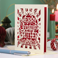 A laser cut cream and dark red Christmas card reading 'MERRY Christmas TO MY BROTHER'. The brother Christmas card is displayed on a tabletop with a blue pen in the foreground and a lit candle in the background, creating a cosy holiday atmosphere.
