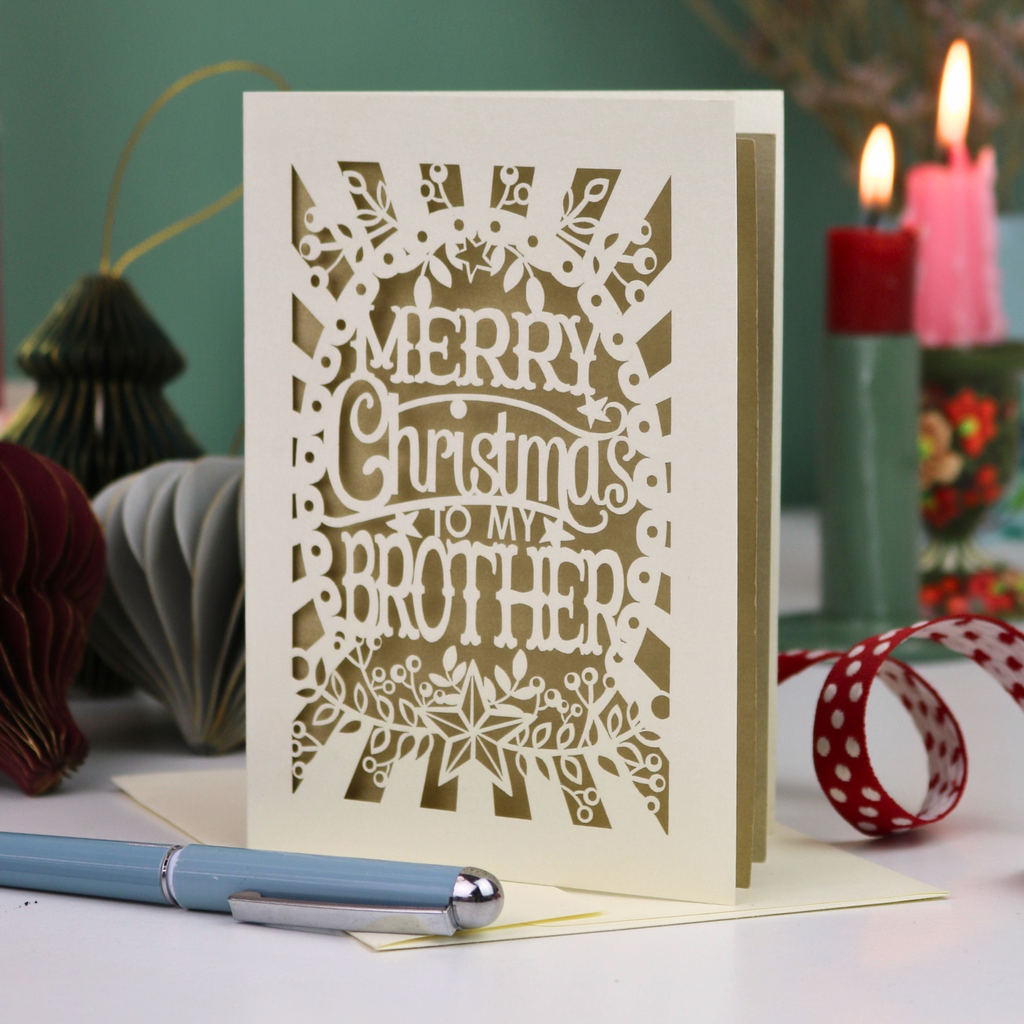 A handmade brother Christmas card reading 'MERRY Christmas TO MY BROTHER'. The card is displayed on a tabletop with a blue pen in the foreground and a lit candle in the background, creating a cosy holiday atmosphere.