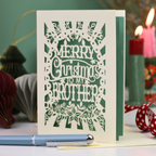 A cream Christmas card  for brother, with intricate laser-cut snowflake and holly designs, featuring the text 'MERRY Christmas TO MY BROTHER'. The card is displayed on a tabletop with a light blue pen in the foreground and festive decorations, including lit candles and a red polka-dot ribbon, blurred in the background.