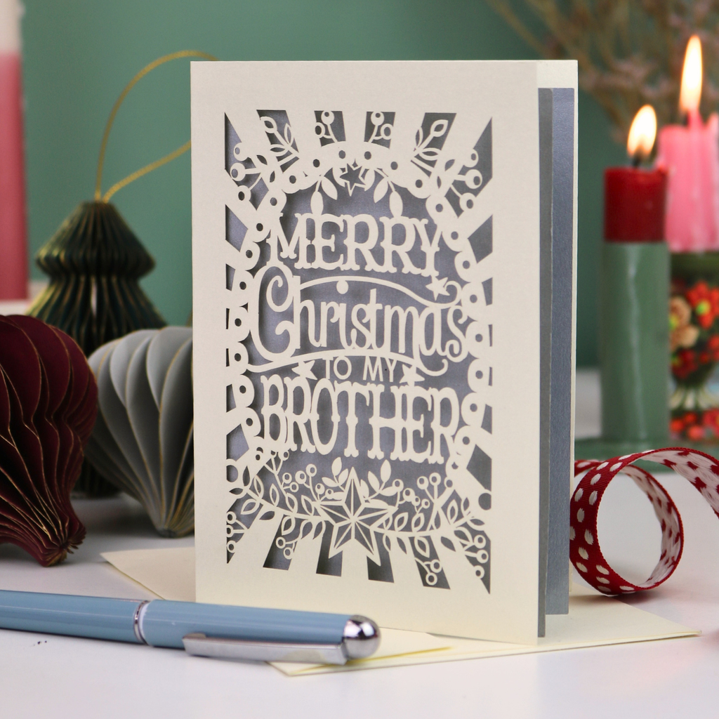 A paper cut Christmas card for brothers, reading 'MERRY Christmas TO MY BROTHER'. The card is displayed on a tabletop with a blue pen in the foreground and a lit candle in the background, creating a cosy holiday atmosphere.