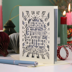 A paper cut Christmas card for brothers, reading 'MERRY Christmas TO MY BROTHER'. The card is displayed on a tabletop with a blue pen in the foreground and a lit candle in the background, creating a cosy holiday atmosphere.