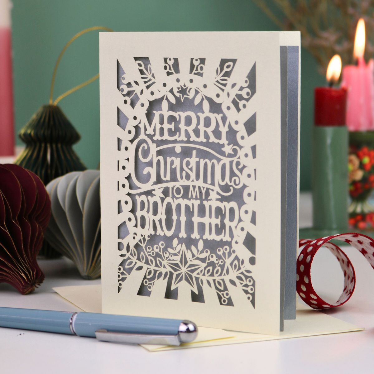 A paper cut Christmas card for brothers, reading 'MERRY Christmas TO MY BROTHER'. The card is displayed on a tabletop with a blue pen in the foreground and a lit candle in the background, creating a cosy holiday atmosphere.