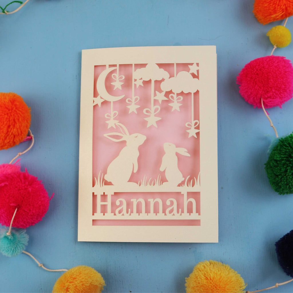 Decorative birthday card with cut-out rabbits and 'Hannah' text on a blue background with colourful pom-poms.
