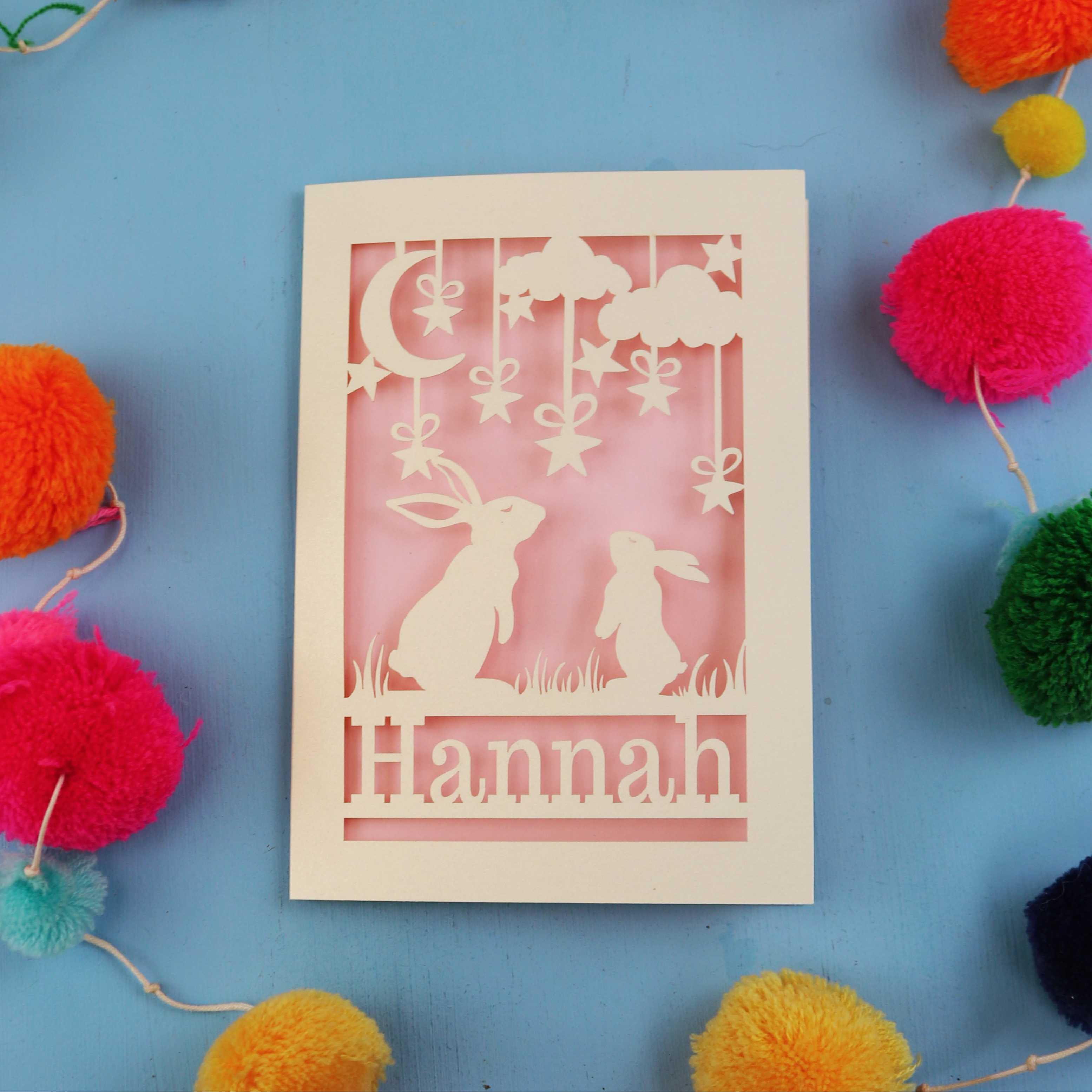 Decorative birthday card with cut-out rabbits and 'Hannah' text on a blue background with colourful pom-poms.