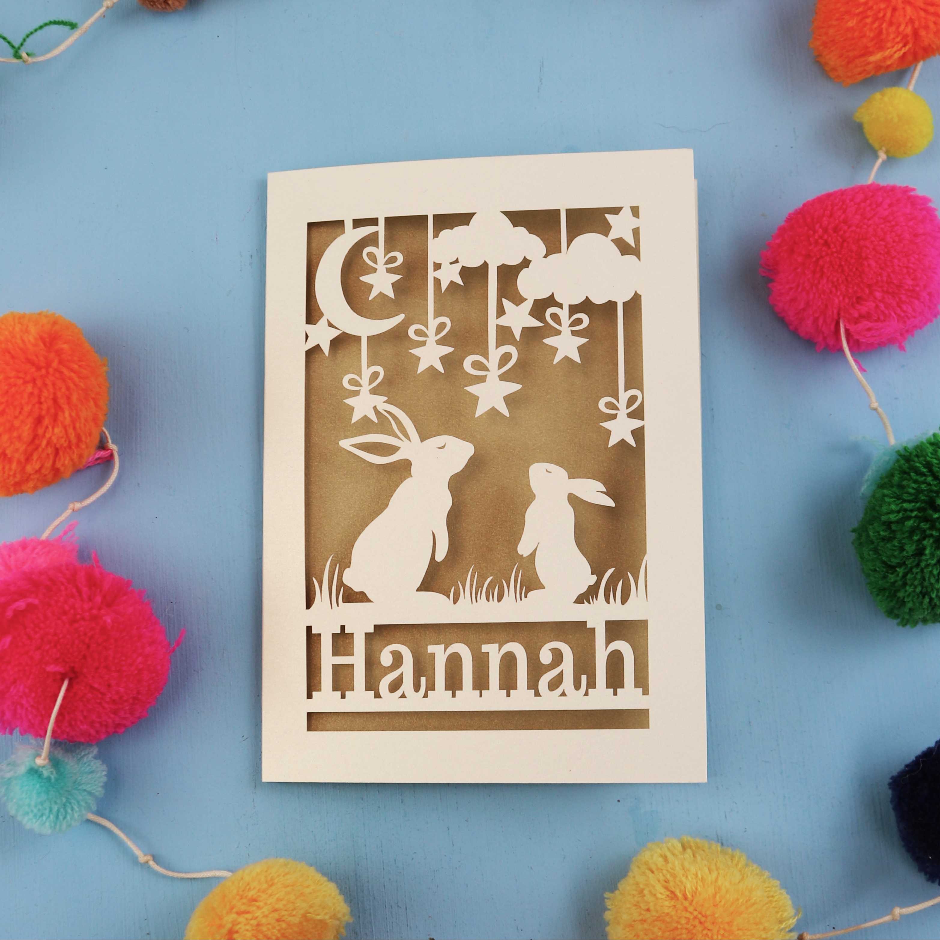 Decorative birthday card with cut-out rabbits and 'Hannah' text on a blue background with colourful pom-poms.