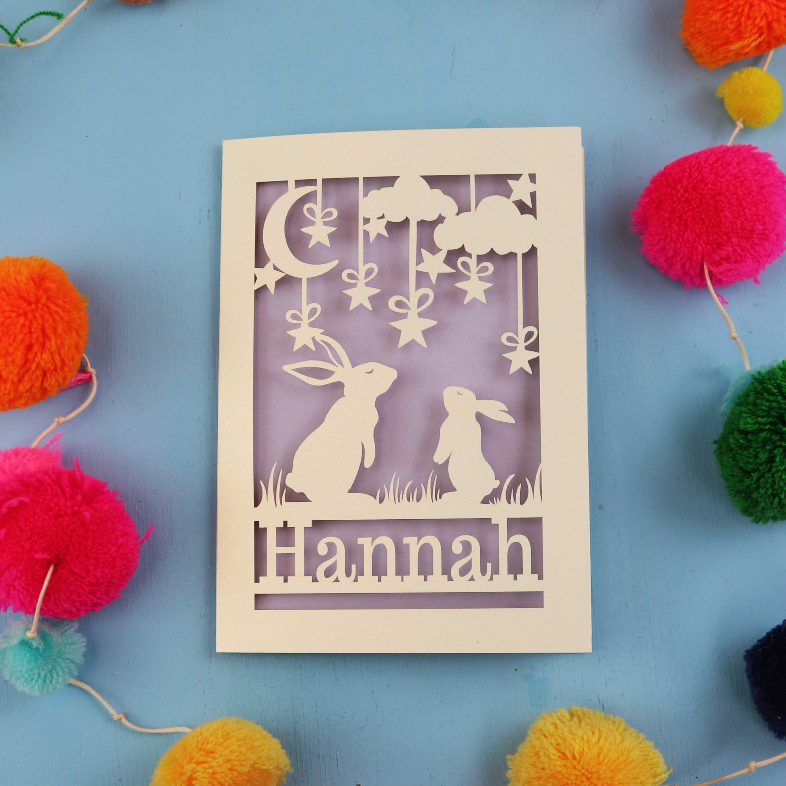 Decorative birthday card with cut-out rabbits and 'Hannah' text on a blue background with colourful pom-poms.
