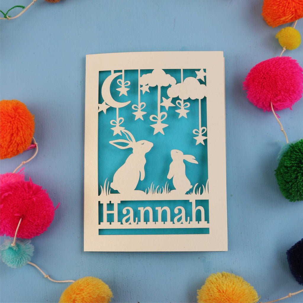 Decorative birthday card with cut-out rabbits and 'Hannah' text on a blue background with colourful pom-poms.