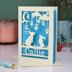 Paper cut birthday Card with cut-out rabbits and 'Callie' text on a blue background, with a pen and decorative items in the background.