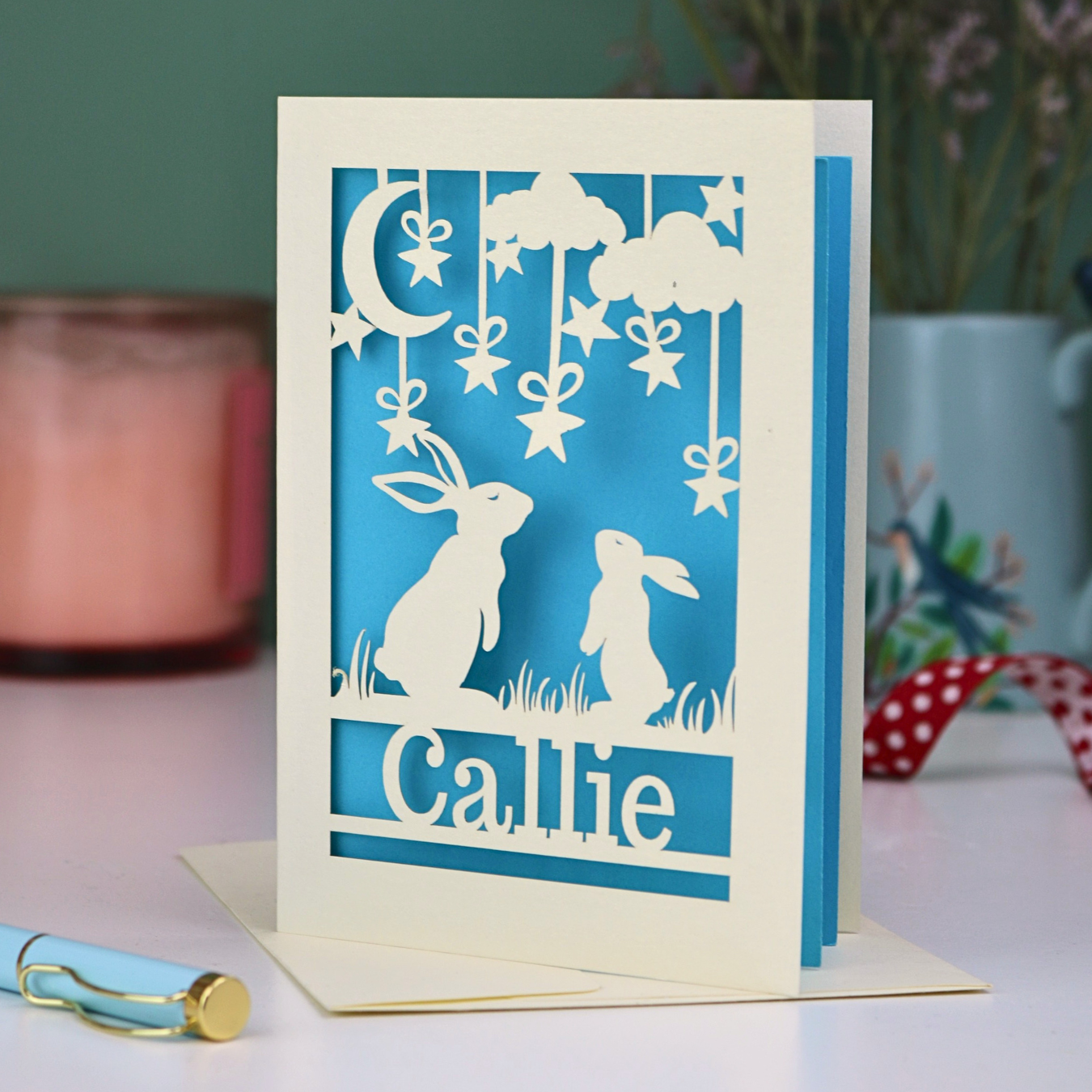 Paper cut birthday Card with cut-out rabbits and 'Callie' text on a blue background, with a pen and decorative items in the background.