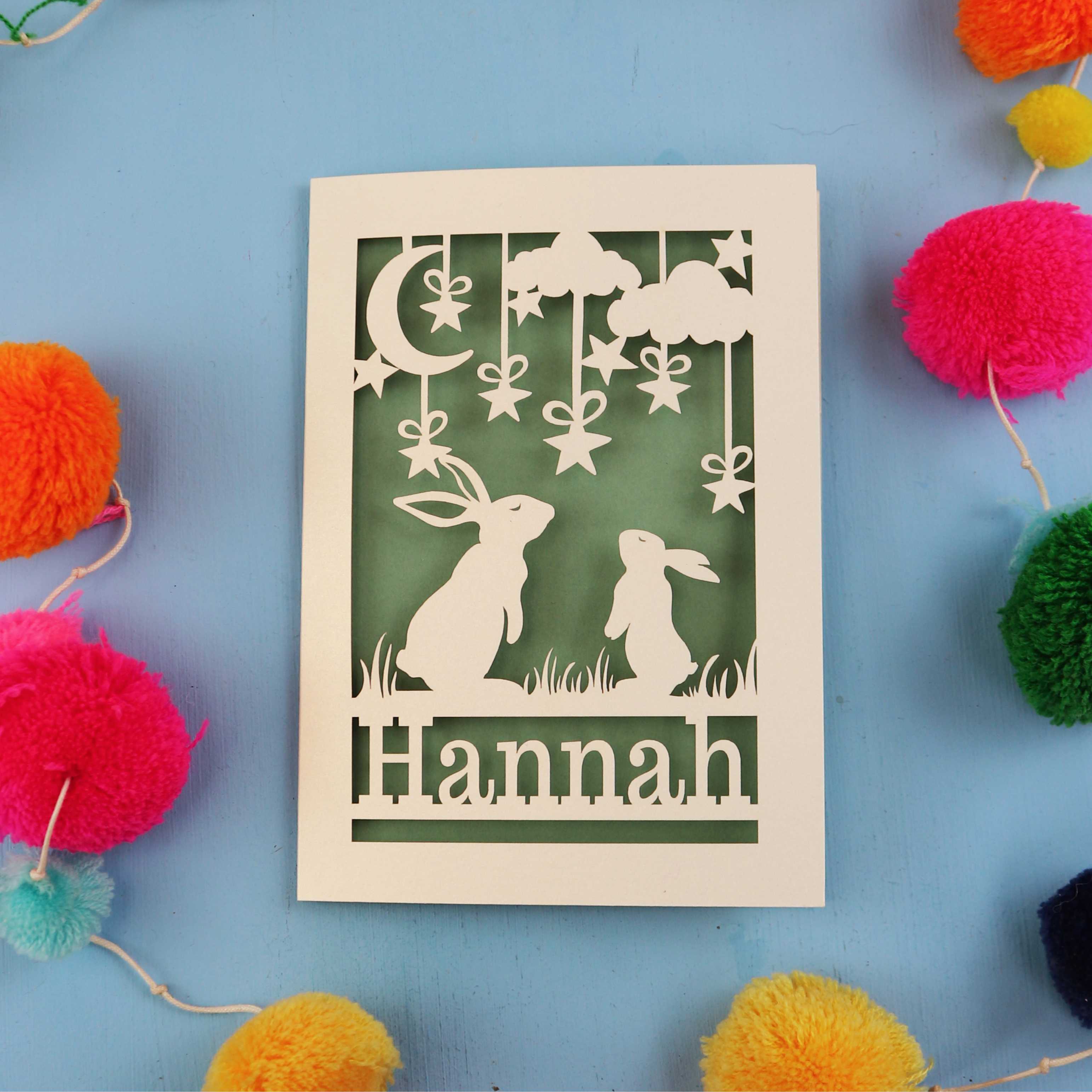 Decorative birthday card with cut-out rabbits and 'Hannah' text on a blue background with colourful pom-poms.