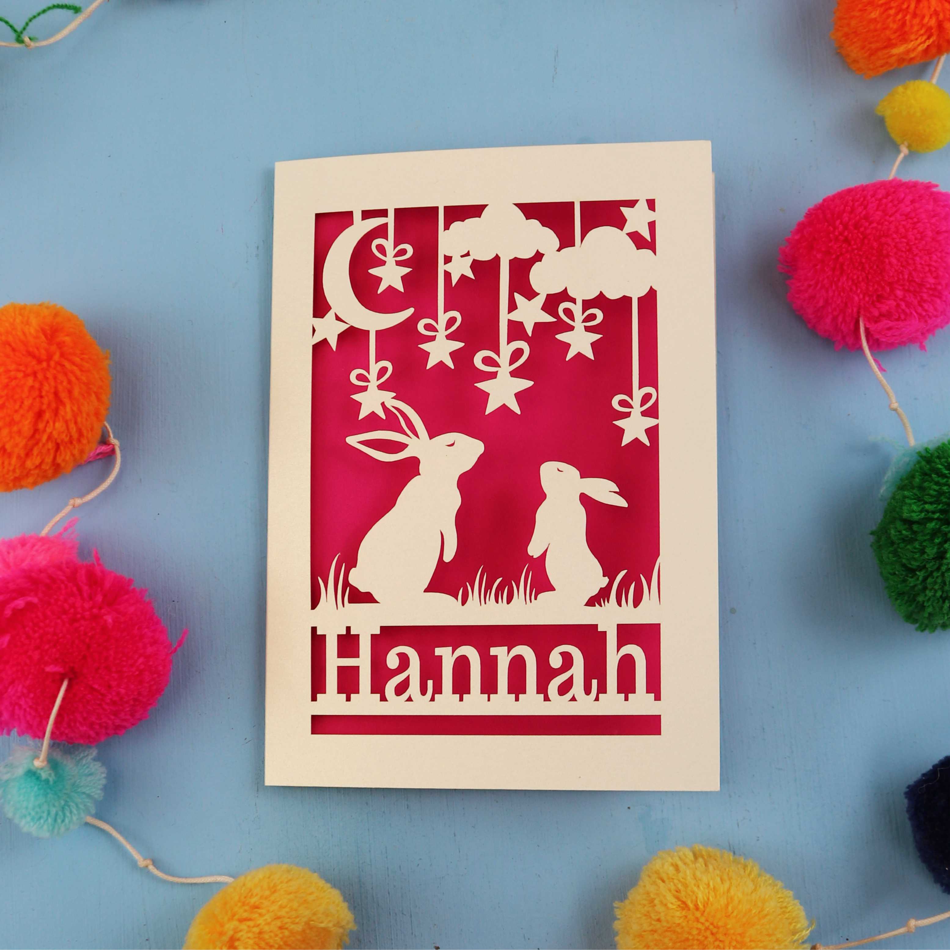Decorative birthday card with cut-out rabbits and 'Hannah' text on a blue background with colourful pom-poms.