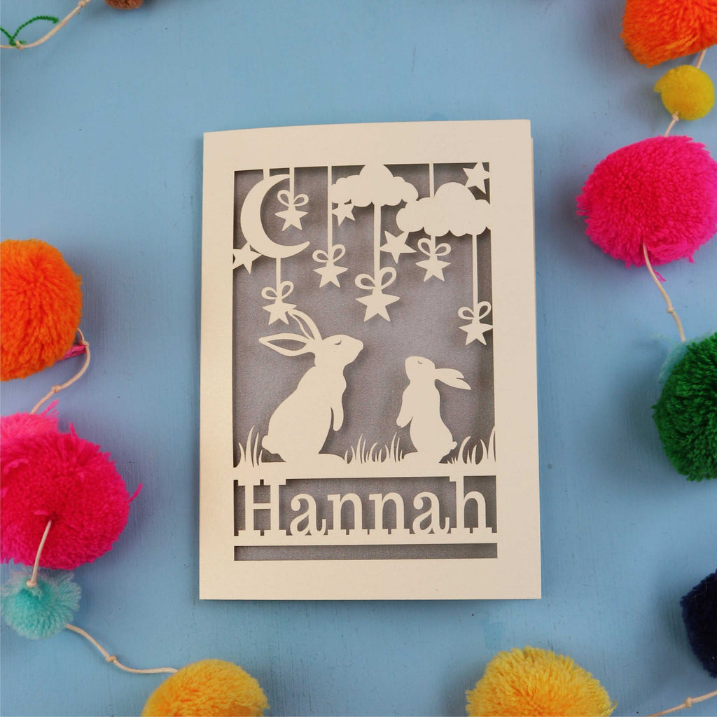 Decorative birthday card with cut-out rabbits and 'Hannah' text on a blue background with colourful pom-poms.