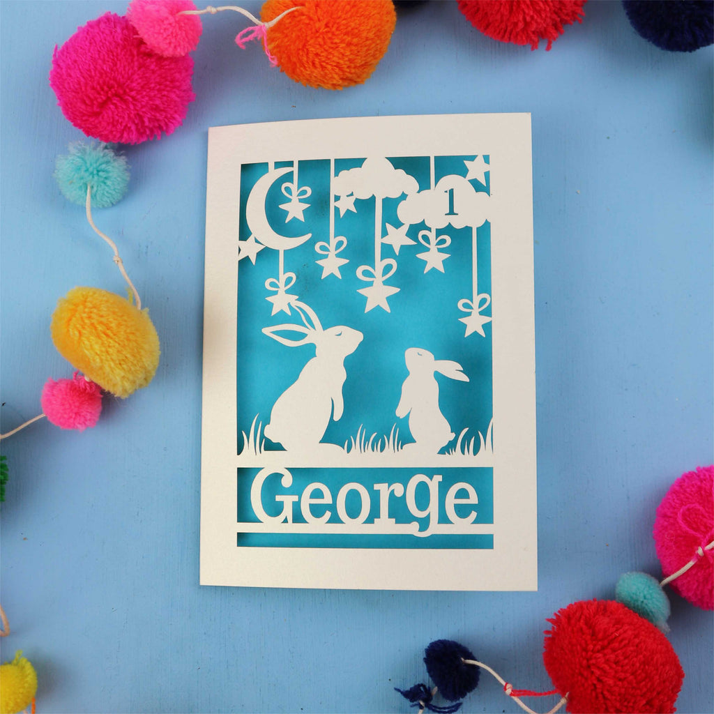 Decorative paper cut out 1st birthday card with two rabbits and the name 'George' on a blue background, surrounded by colourful pom-poms.