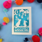 Decorative paper cut out 1st birthday card with two rabbits and the name 'George' on a blue background, surrounded by colourful pom-poms.