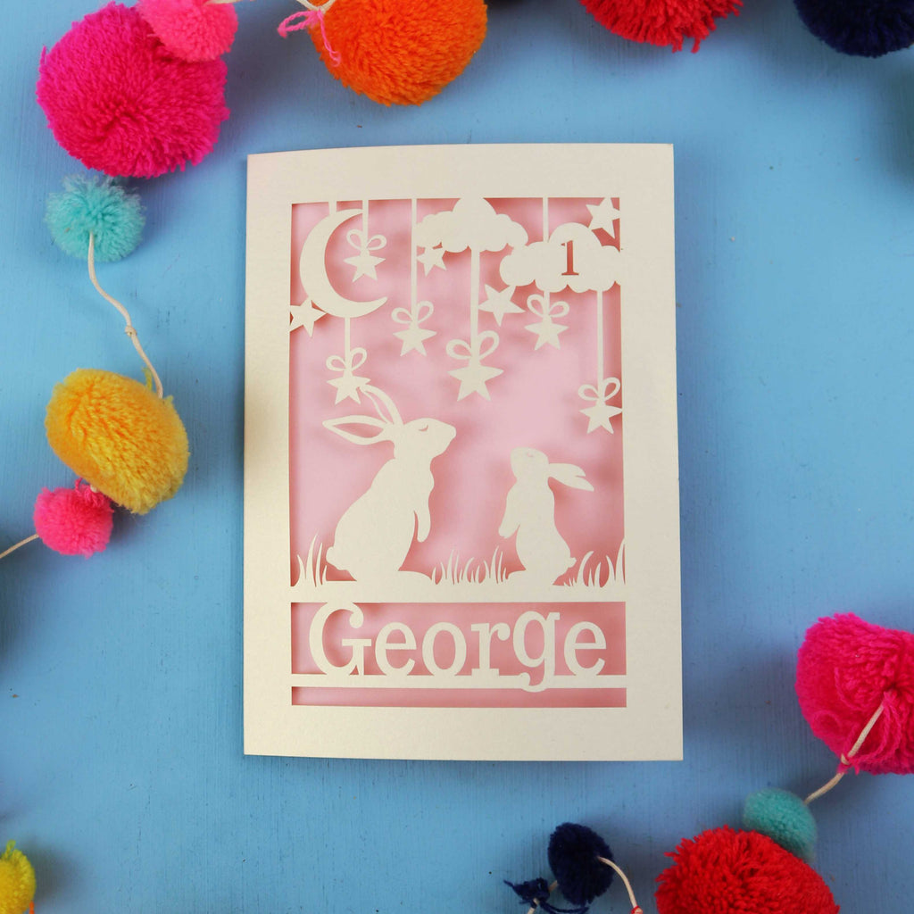 Decorative laser cut first birthday card with two rabbits and the name 'George' on a blue background, surrounded by colourful pom-poms.