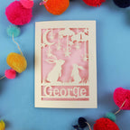 Decorative laser cut first birthday card with two rabbits and the name 'George' on a blue background, surrounded by colourful pom-poms.
