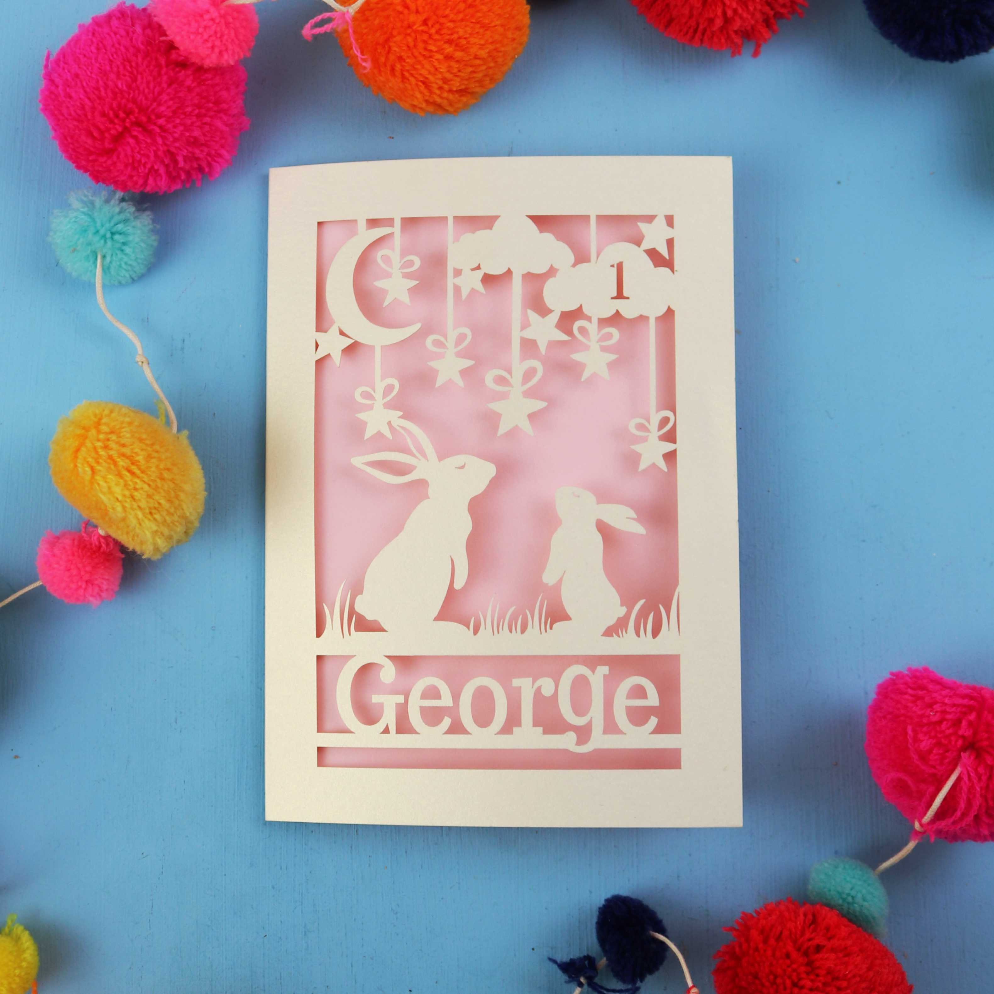 Decorative laser cut first birthday card with two rabbits and the name 'George' on a blue background, surrounded by colourful pom-poms.
