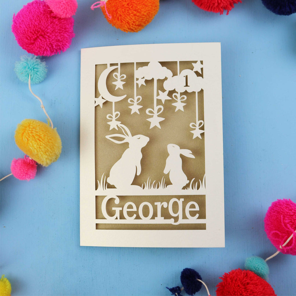 Decorative laser cut 1st birthday card with two rabbits and the name 'George' on a blue background, surrounded by colourful pom-poms.