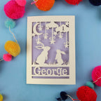 Decorative laser cut 1st birthday card with two rabbits and the name 'George' on a blue background, surrounded by colourful pom-poms.