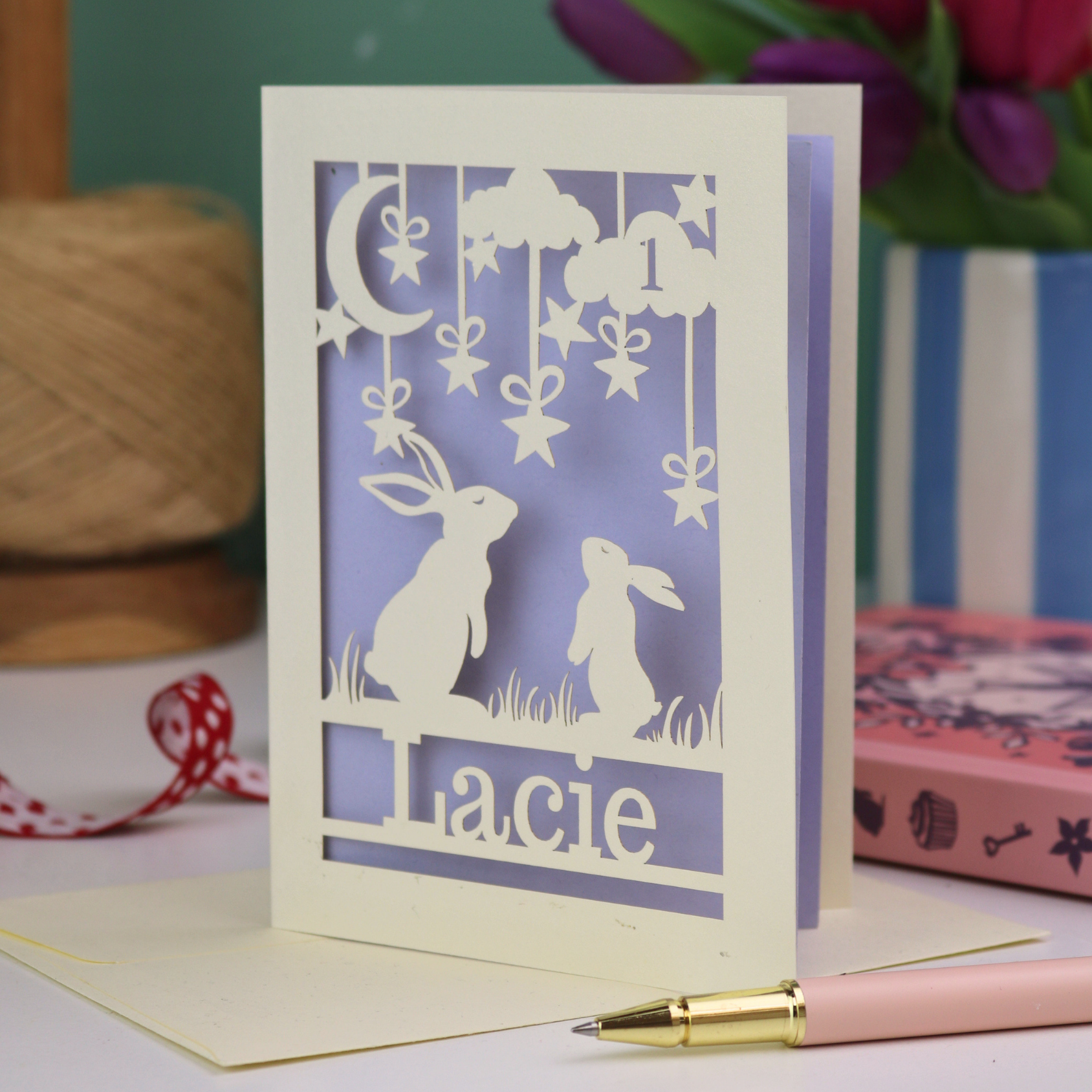 A laser cut 1st birthday card with cut out bunnies looking up at the moon and stars, personalised with the age 1 in a cloud in the sky, and the name "Lacie" at the bottom. 