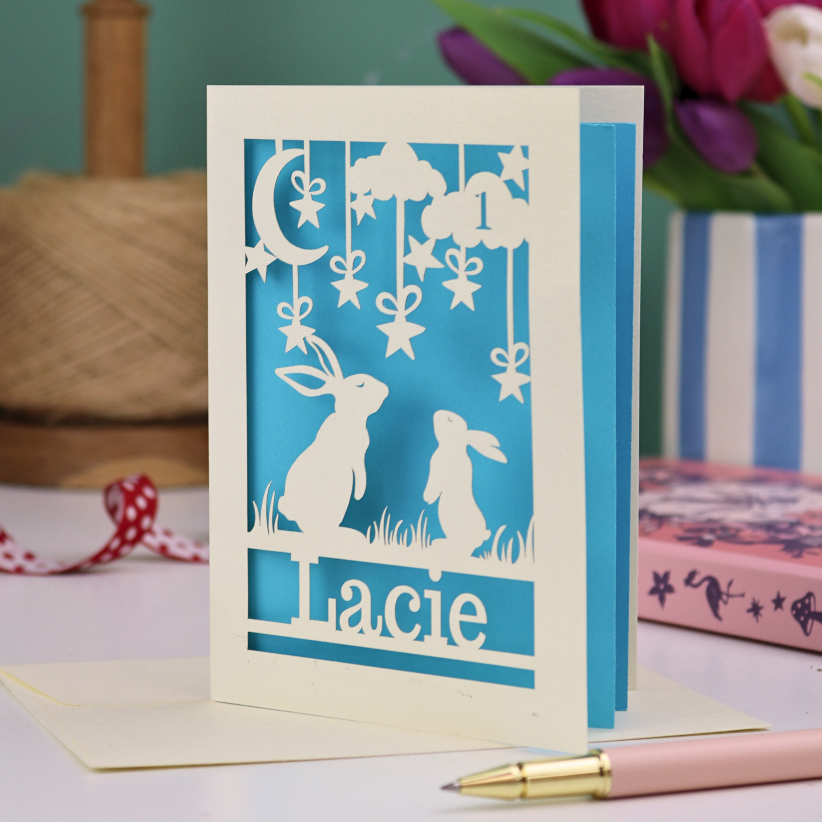 A laser cut 1st birthday card with cut out bunnies looking up at the moon and stars, personalised with the age 1 in a cloud in the sky, and the name "Lacie" at the bottom. 