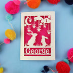 Decorative laser cut 1st birthday card with two rabbits and the name 'George' on a blue background, surrounded by colourful pom-poms.