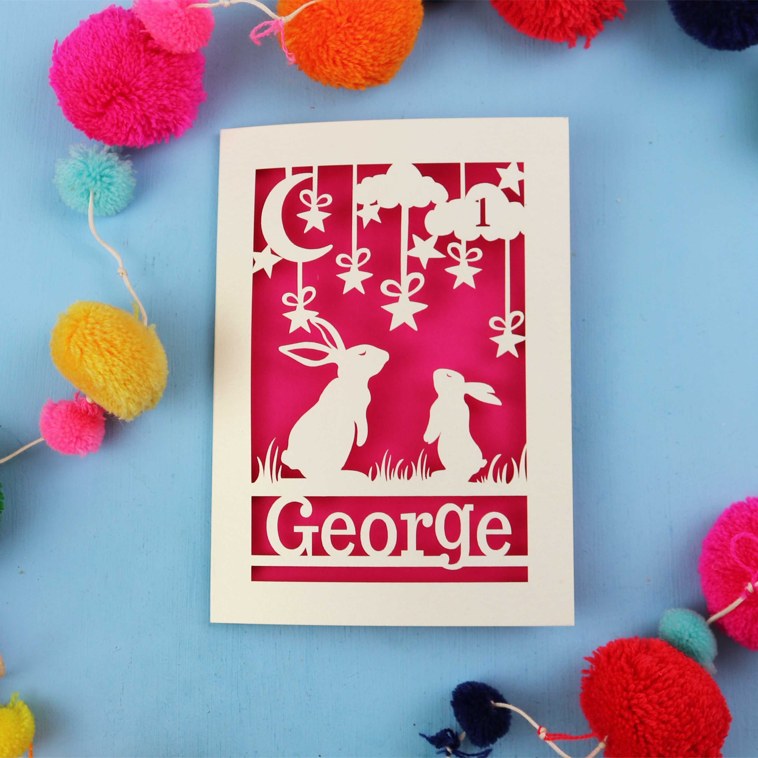 Decorative laser cut 1st birthday card with two rabbits and the name 'George' on a blue background, surrounded by colourful pom-poms.