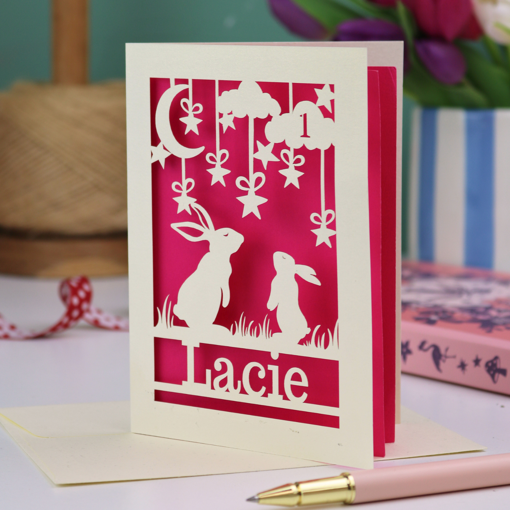 A laser cut 1st birthday card with cut out bunnies looking up at the moon and stars, personalised with the age 1 in a cloud in the sky, and the name "Lacie" at the bottom. 