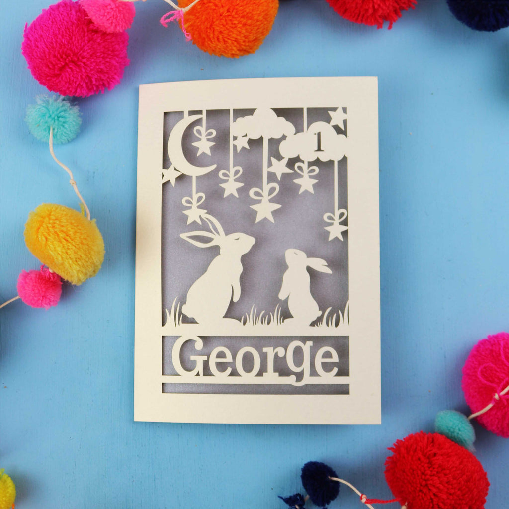 Decorative laser cut 1st birthday card with two rabbits and the name 'George' on a blue background, surrounded by colourful pom-poms.