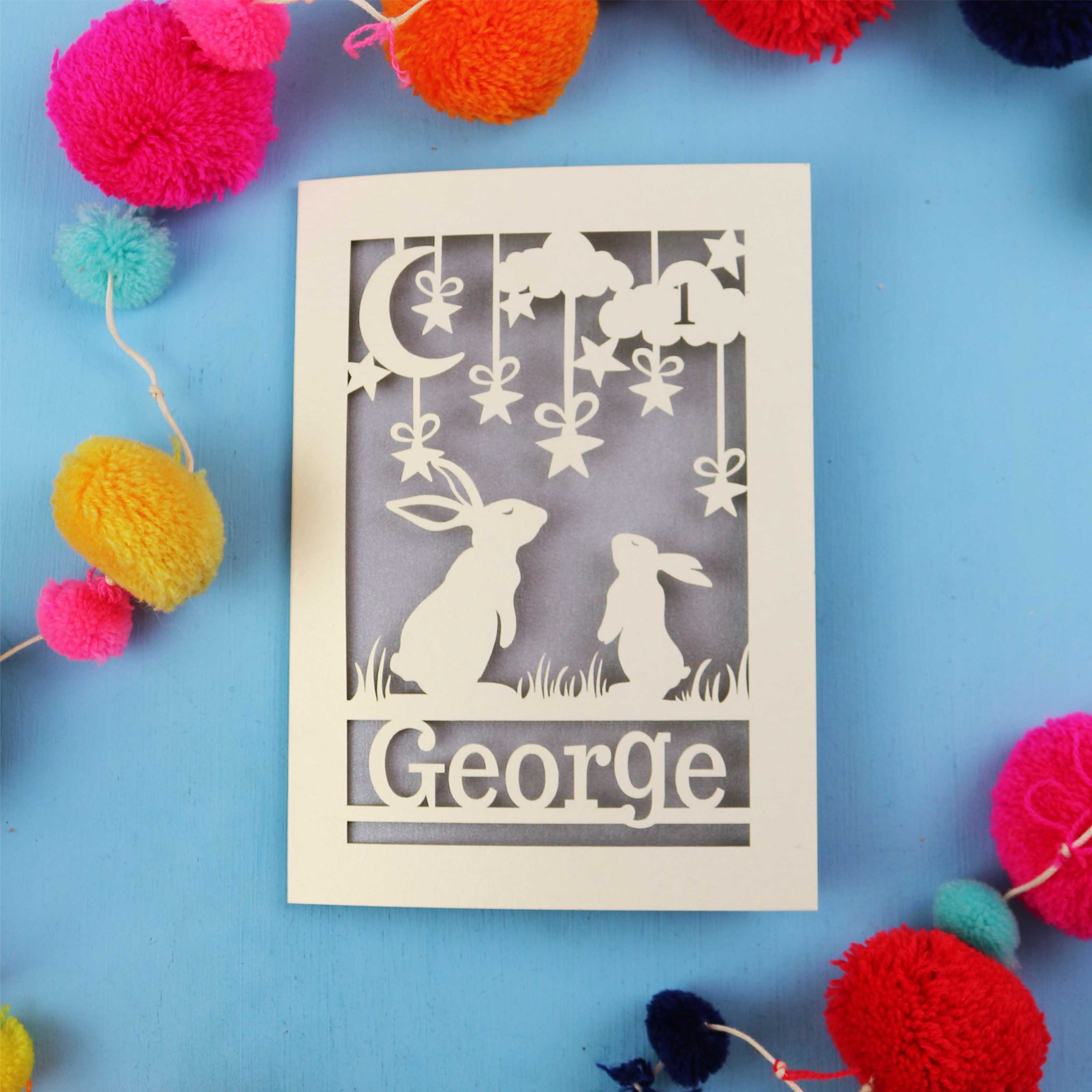Decorative laser cut 1st birthday card with two rabbits and the name 'George' on a blue background, surrounded by colourful pom-poms.