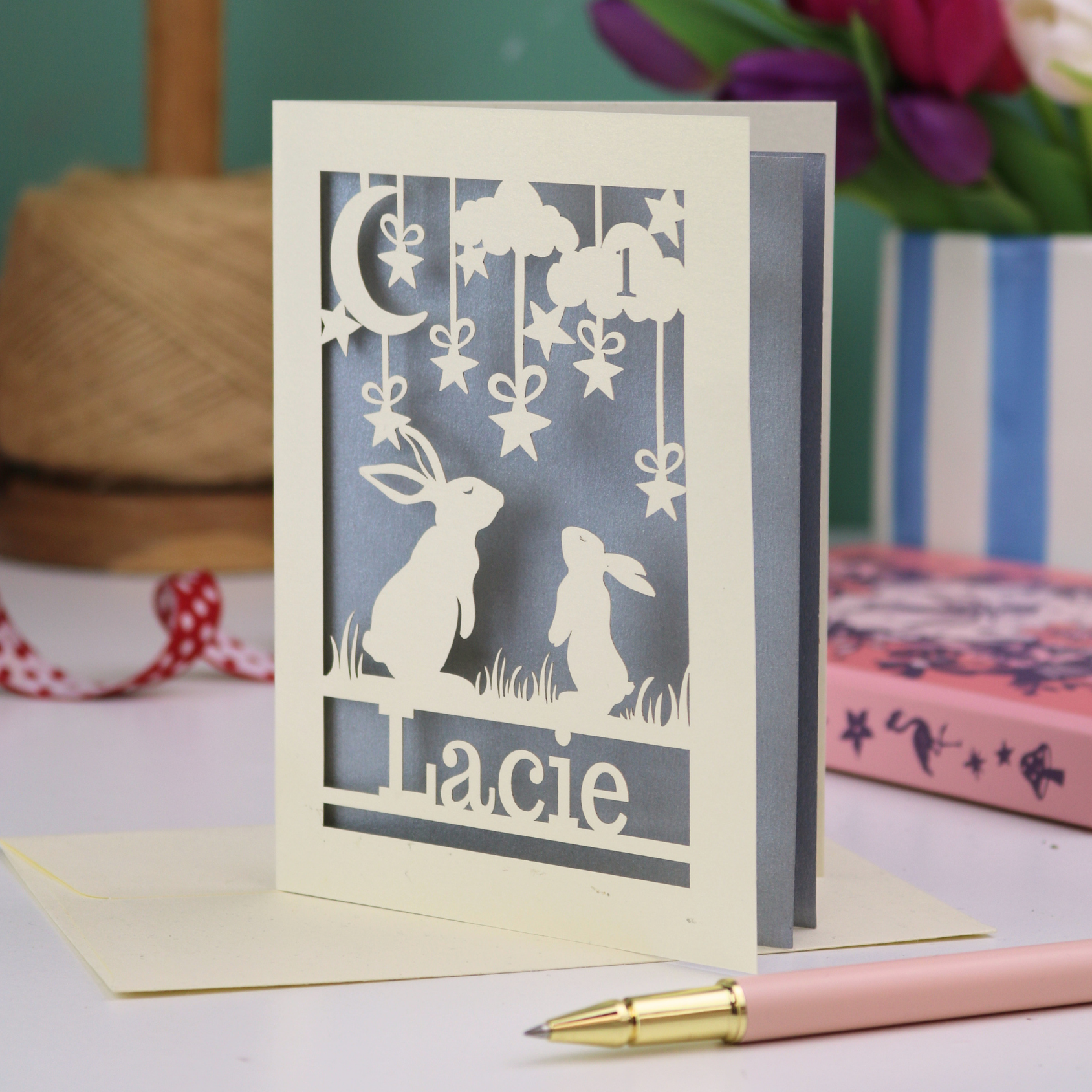 A laser cut 1st birthday card with cut out bunnies looking up at the moon and stars, personalised with the age 1 in a cloud in the sky, and the name "Lacie" at the bottom. 
