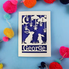 Decorative laser cut 1st birthday card with two rabbits and the name 'George' on a blue background, surrounded by colourful pom-poms.